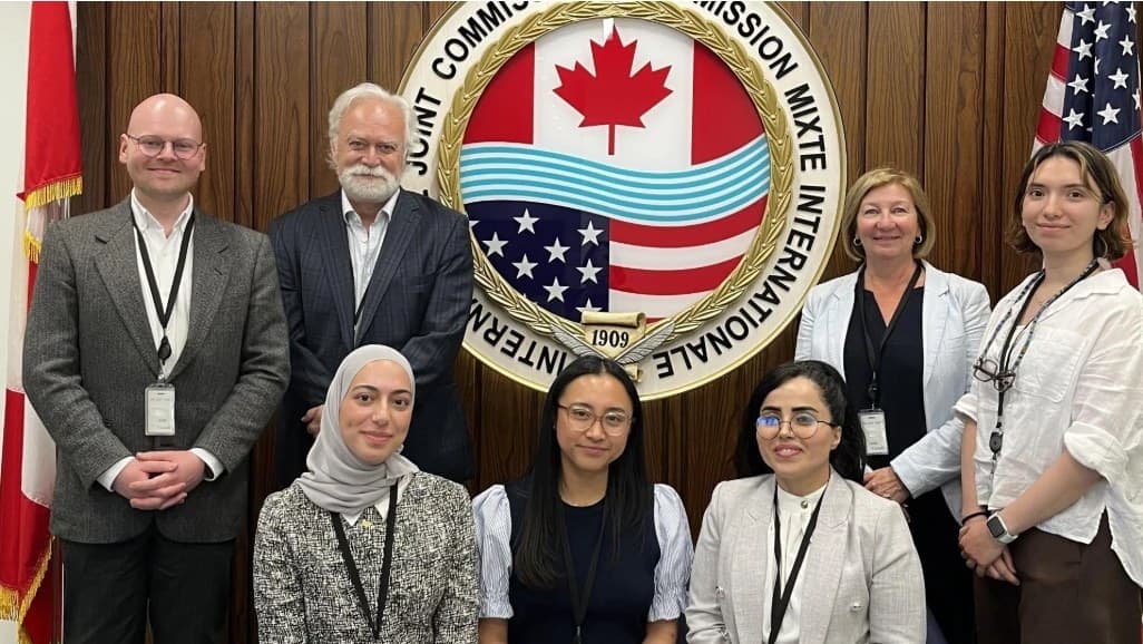Seven people, students and staff, pose in front of the International Joint Commission’s seal, between the Canadian and American flags.