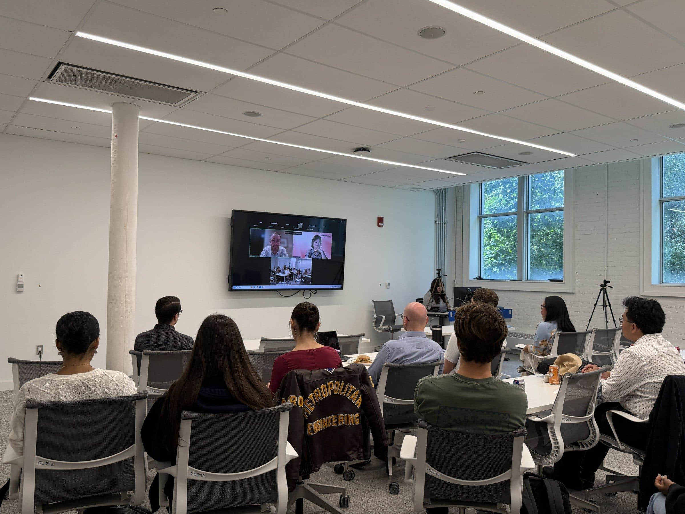Attendees at tables in a bright seminar room watch a wall screen where both speakers appear on a video call.